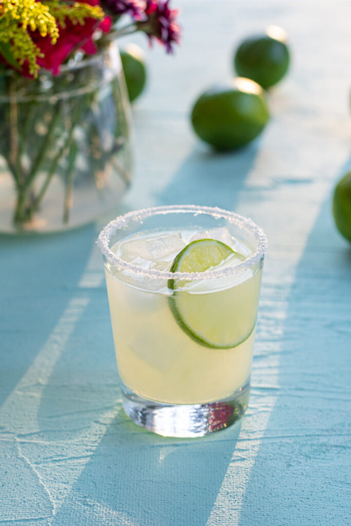 A glass with a salt rim filled with classic margarita cocktail and ice, topped with a lime wheel, in the background limes and flowers. 