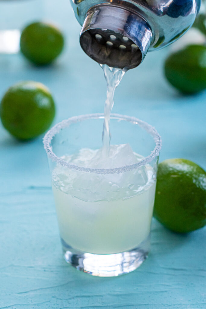 A cocktail shaker pouring margarita into a glass filled with ice.